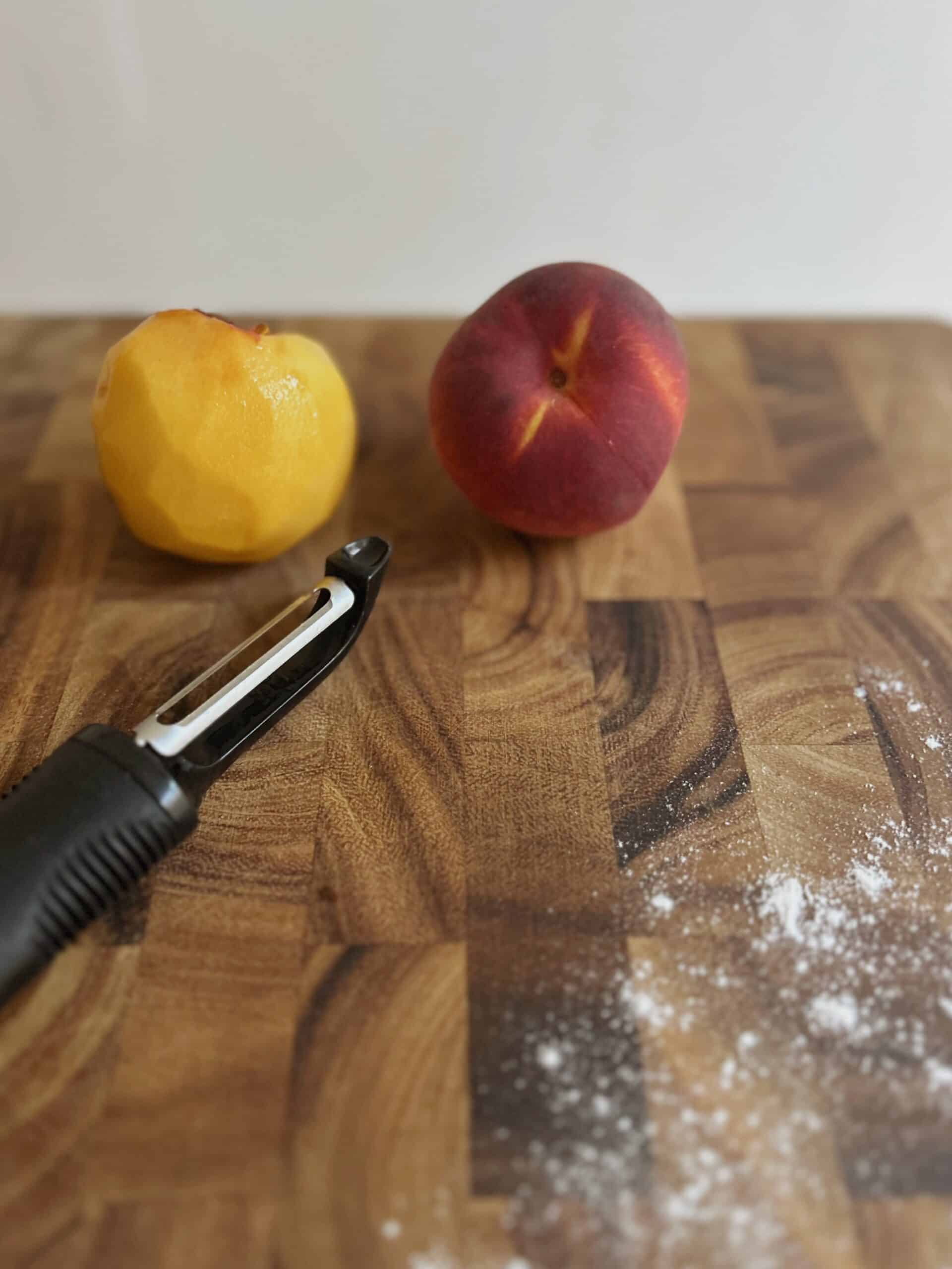 Peeled peach and a peach with the skin on pictured with a peeler next to it.